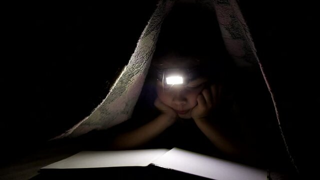 little girl is reading a book under a blanket with a flashlight in a dark room at night