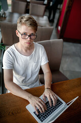 Young guy in a white t-shirt and glasses works on a laptop sitting at a table in a cafeteria
