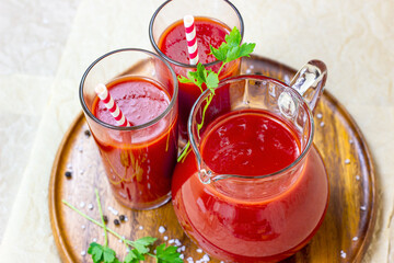 Top view of fresh red tomato juice in a glass with a straw and jar on light wooden background.