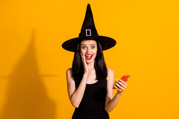 Photo of pretty lovely young magician lady hold telephone hand cheek open mouth excited find online shop discount wear black wizard headwear dress isolated bright yellow color background