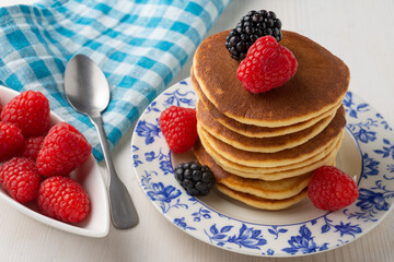 Close-up of plate with pancakes, spoon, blackberries and raspberries, on white wooden table with blue checkered cloth and bowl with raspberries, horizontal