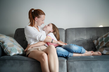 Obraz premium Young beautiful mother and her daughter child 8 years old sitting on a gray sofa and tenderly hugging, happiness and love in the family. Mother's day concept