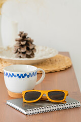 Obraz na płótnie Canvas a white mug with a blue pattern, a Notepad and sunglasses on a brown wooden table on a white background. Business lunch. Scandinavian style.