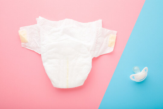White baby diaper and soother on light pink blue table background. Pastel color. Closeup. Top down view.