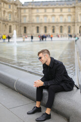 Young man wearing black suit sitting near fountain and smoking in Paris, France.