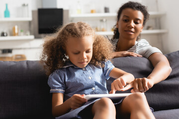 selective focus of african american nanny pointing with finger at digital tablet in hands of child sitting on sofa