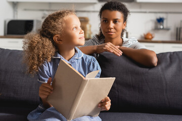 selective focus of african american nanny behind dreamy child looking away while holding open book