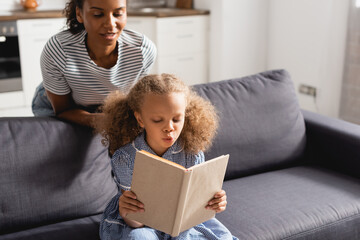 african american nanny in striped t-shirt looking at concentrated girl sitting on couch and reading book