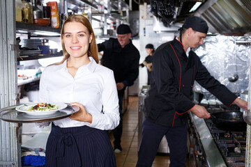 Young positive waitress with tray standing at restaurant kitchen among kitchen workers