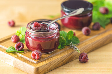 Two glass jar of fresh homemade raspberry jam on a wooden background. Several fresh berries and mint are near it.