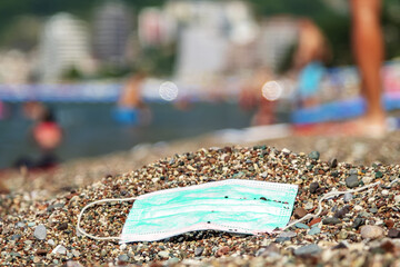 Discarded used face mask lies on a pebble beach, beachgoers are relaxing by the sea in the background. Becici, Budva Municipality, Montenegro