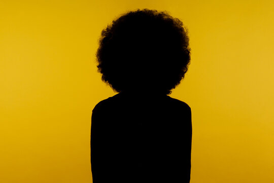 No name, anonymous hiding face in shadow, human identity. Silhouette portrait of curly hair person standing calm alone in darkness with hands down. indoor studio shot isolated on yellow background.