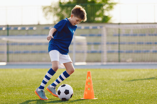 Boy in soccer uniform practice with ball. Child kicking ball on grass stadium. Young athlete improving football dribbling skills on training field. Kid soccer player dribbling through cones