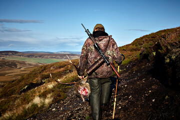A hunter walking in the Scottish highlands with a dead deer trophy in his hand.