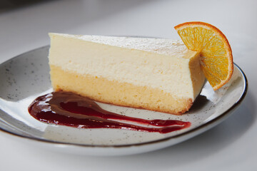 A slice of cheesecake with raspberry sauce decorated with dried orange piece. Dessert served on the white plate at the restaurant.