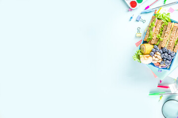 Healthy children school lunch box: sandwich, vegetables ,fruit and mineral water bottle, with school supplies on, on light blue background copy space