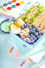 Healthy children school lunch box: sandwich, vegetables ,fruit and mineral water bottle, with school supplies on, on light blue background copy space