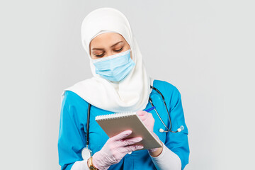 Fototapeta na wymiar friendly muslim doctor or nurse wearing hijab and medical face mask with stethoscope writing prescription to patient on gray background