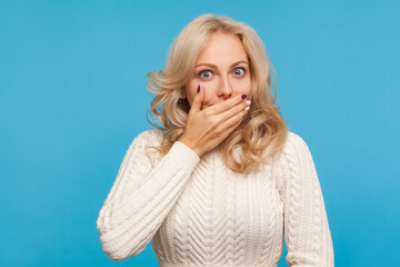 Closeup scared shocked blond woman closing mouth with hand, afraid to say too much, embarrassment. Indoor studio shot isolated on blue background