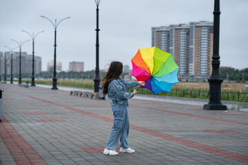 The girl in a denim jacket and jeans with effort holds an umbrella from wind. Strong wind pulls out a rainbow umbrella from her hands. Wind has disheveled hair