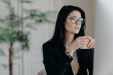 Photo of concentrated brunette female copywriter drinks coffee, works on creative task, wears...