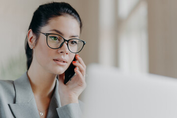 Close up shot of pleasant looking brunette European woman wears transparent glasses, focused in laptop screen, has telephone conversation during remote work, works on project. Technology concept