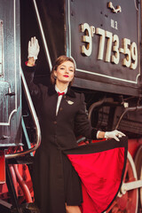 A young lady in vintage clothes standing on stairs of a historical retro train and waving her hand