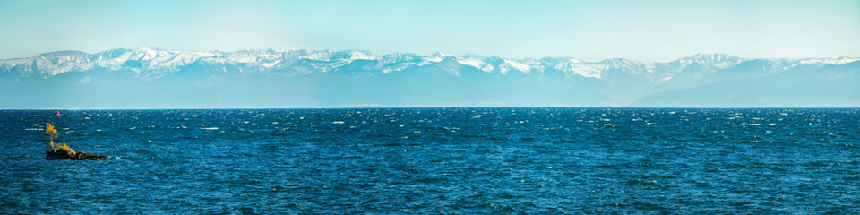 Panorama of blue water of Lake Baikal, with mountain peaks against the sky with a tree in the middle of summer