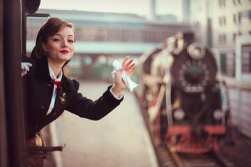 A young beautiful lady waving with het white glove from a window of a train; a historical retro locomotive is seen on the background