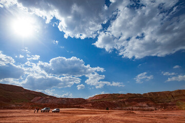 Fototapeta na wymiar Red bauxite clay. Aluminium ore mining. Quarry of red ground on blue sky with clouds. Two off-road cars and two people.