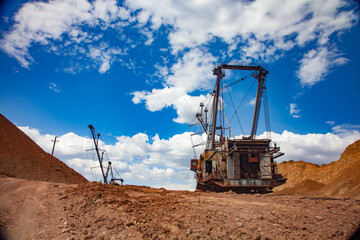Fototapeta premium Aluminium ore quarry. Bauxite clay open-cut mining. Walking dragline excavators and empty rocks. On red and orange soil and blue sky with clouds.