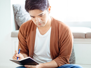 Cross-legged young asian man writing in notebook.