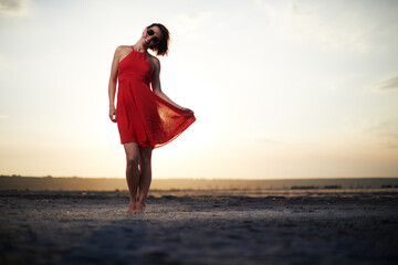 Brunette adult woman in red dress and sunglasses posing in salt lake on beautiful sunset background