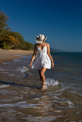 A beautiful young curly-hair woman in a little white/beige dress and cowboy's straw hat  is posing at the beach. Portrait Photography.