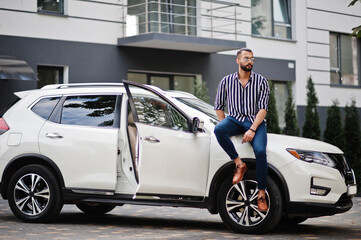 Successful arab man wear in striped shirt and sunglasses sitting on the hood of white suv car. Stylish arabian men in transport.