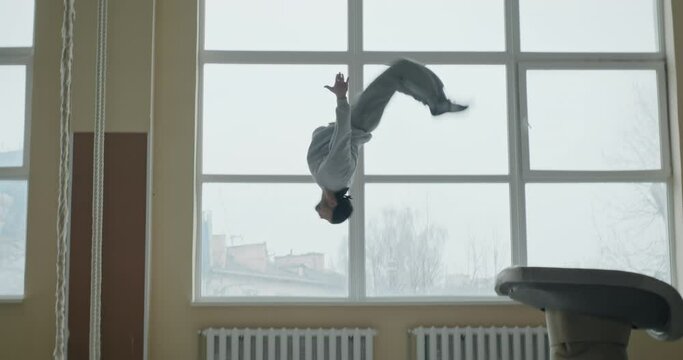 Side view of young male gymnast jumping from vault and performing back flip in slow motion during training in sports hall
