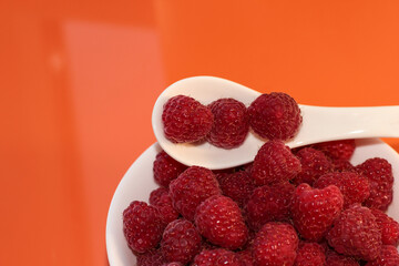 Ripe raspberries in a white plate on an orange background. On a white spoon raspberries.