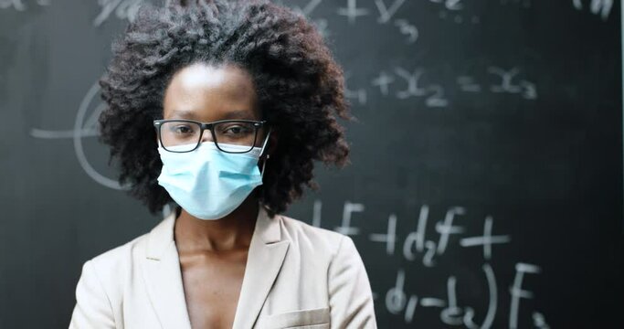 Portrait of young African American female teacher in glasses and medical mask looking at camera in classrom. Blackboard with formulas on background. Coronavirus concept. Pandemic schooling.