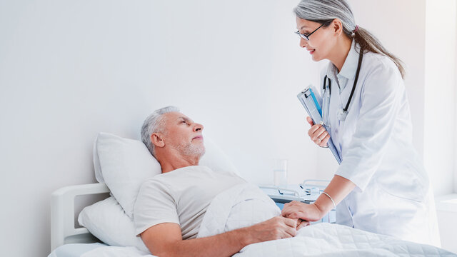 Smiling Middle Aged Doctor With Clipboard Attending Sick Man In Hospital Ward