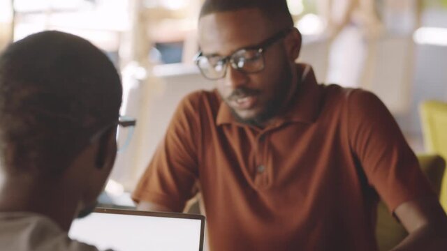 Over the shoulder shot of young cheerful afro-american man greeting colleague with fist bump, sitting at cafe table and discussing business on lunch