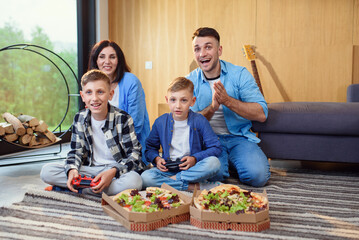 Happy stylish family sitting on floor, playing video games with gamepads and eating tasty pizza.