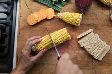 Fototapeta na wymiar Female hand cutting fresh corn cob in half on a wooden cutting board