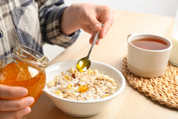 Woman putting honey into bowl with muesli at wooden table, closeup