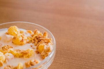 Crunches and corn flakes with yogurt in a transparent glass bowl for cooking in the morning at home on a wooden table in the sunlight, mockup.