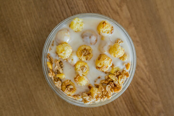 Crunches and corn flakes with yogurt in a transparent glass bowl for cooking in the morning at home on a wooden table in the sunlight, top view.