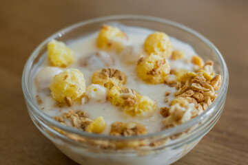 Crunches and corn flakes with yogurt in a transparent glass bowl for cooking in the morning at home on a wooden table in the sunlight.