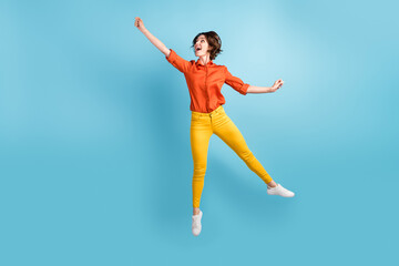 Fototapeta na wymiar Full body photo of attractive funny lady jump high flight raising up air imaginary umbrella rejoicing weekend wear orange shirt yellow trousers sneakers isolated blue color background