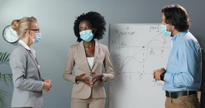 Mixed-races women and man in medical msks discussing study concept in classroom with physics or math formulas board on background. Multiethnic male and females talking in office about business growing