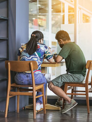 Asian couple customers wear face mask using smartphone while sit in waiting social distance queue for take away drink in coffee shop. New normal lifestyle concept. Selective focus on male.