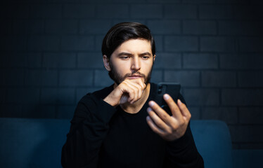Portrait of young thoughtful man, looking in smartphone, sitting on sofa. Background of black brick wall.